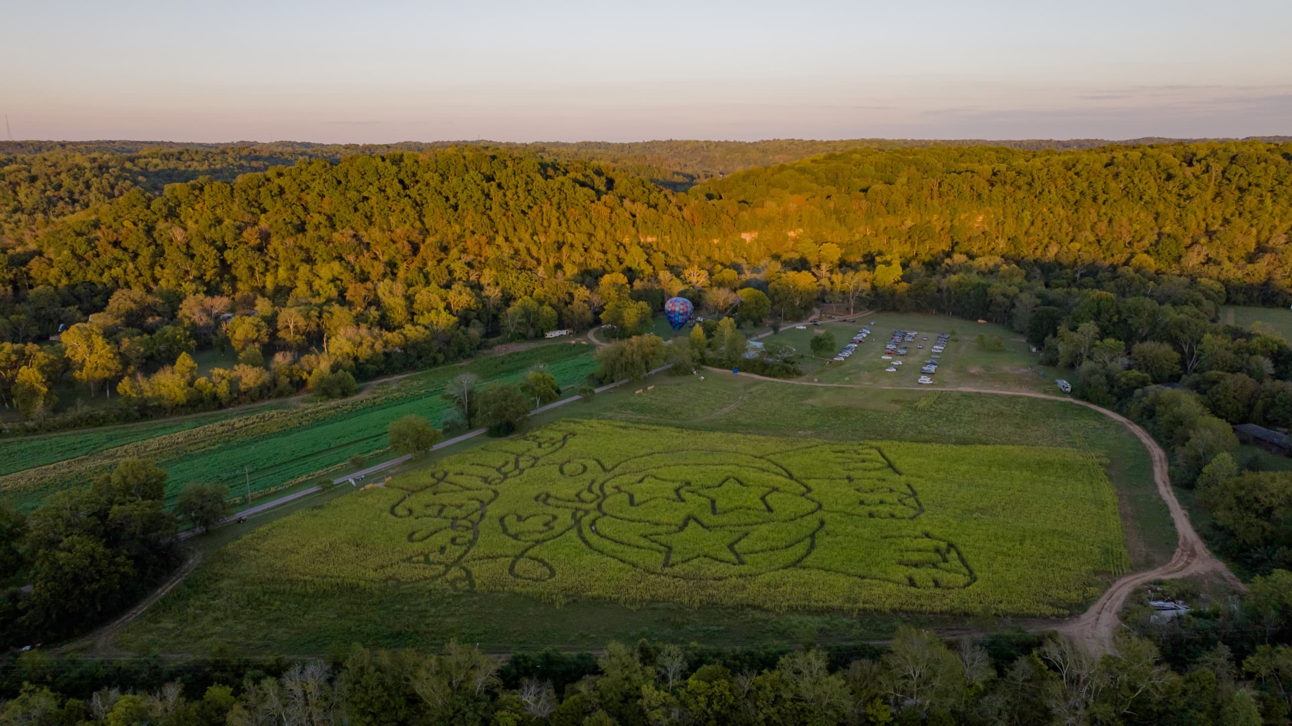 Golden-hour aerial of the Nashville Fall Festival — the hillside corn maze, a hot air balloon, and the wooded South Harpeth River valley in Bellevue
